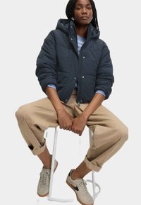 Polstret navy jakke med hætte, frontknaplukning og to lommer, båret over en stribet skjorte. Modellen kombinerer den med beige bukser og sneakers.