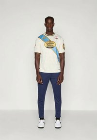 Camisola desportiva branca com uma faixa diagonal azul, detalhes amarelos e logótipo; calças de treino azuis-escuro; sapatilhas brancas com detalhes pretos.