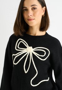 Pullover nero in maglia con un design a fiocco in filato bianco al centro, dotato di un pom-pom testurizzato al nodo del fiocco.