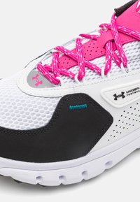Valkoiset urheilukengät mustilla ja vaaleanpunaisilla yksityiskohdilla, vaaleanpunaiset nauhat, verkkomateriaali päällä ja Under Armour -logo sivulla ja kielellä.