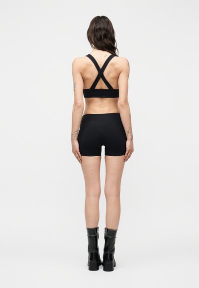Femme debout de dos, portant un soutien-gorge de sport noir à dos croisé, un short taille haute assorti et des bottes noires mi-mollet.