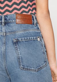 Persona che indossa jeans di denim azzurro chiaro con patch in pelle "Weekend MaxMara" e una maglietta a righe ondulate rosa e nera, mostrando il dettaglio della tasca posteriore.