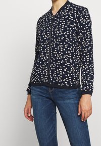 Kvinna klädd i en marinblå zip-up jacka med små vita och beige blommönster samt blå denimjeans mot en enfärgad bakgrund.