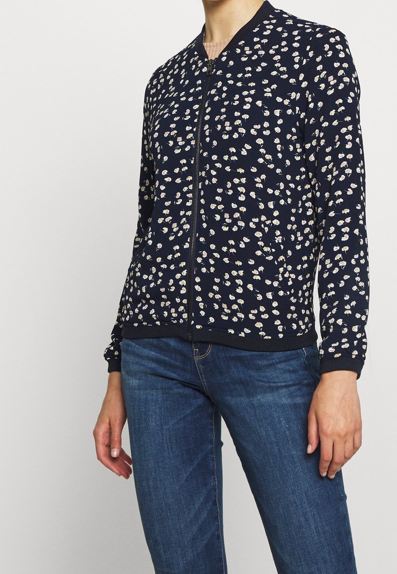 Kvinna klädd i en marinblå zip-up jacka med små vita och beige blommönster samt blå denimjeans mot en enfärgad bakgrund.