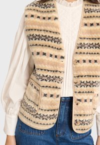 Beige gemusterte Weste aus weichem Stoff, mit horizontalen Streifen in Schwarz und Pink, abgerundeten Kanten und drei Knöpfen auf der Vorderseite.