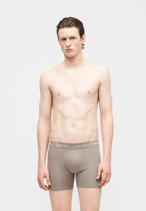 Joven de pie frente a un fondo blanco, vistiendo calzoncillos bóxer beige de Calvin Klein, mirando hacia adelante con expresión neutra.