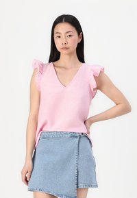 Top rosa senza maniche con scollatura a V e dettagli arricciati sulle spalle, abbinato a una gonna avvolgente di denim con orlo leggermente sfrangiato.