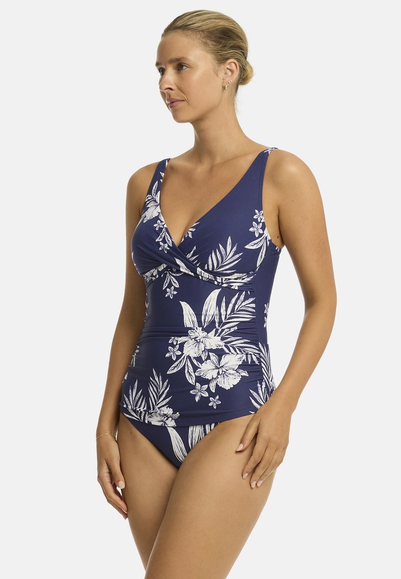 Modelka nosi granatowy kostium kąpielowy z białymi kwiatowymi wzorami, stoi na jednolitym tle, patrzy w bok z delikatnym wyrazem twarzy.