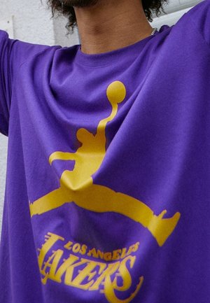 Osoba nosząca fioletową koszulkę z żółtym konturem skaczącego zawodnika koszykówki oraz napisem "Los Angeles Lakers" poniżej.