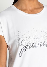 Camiseta de algodón blanca de manga corta, con rhinestones plateados dispuestos en un patrón brillante, con la palabra "sparkle" en negro.