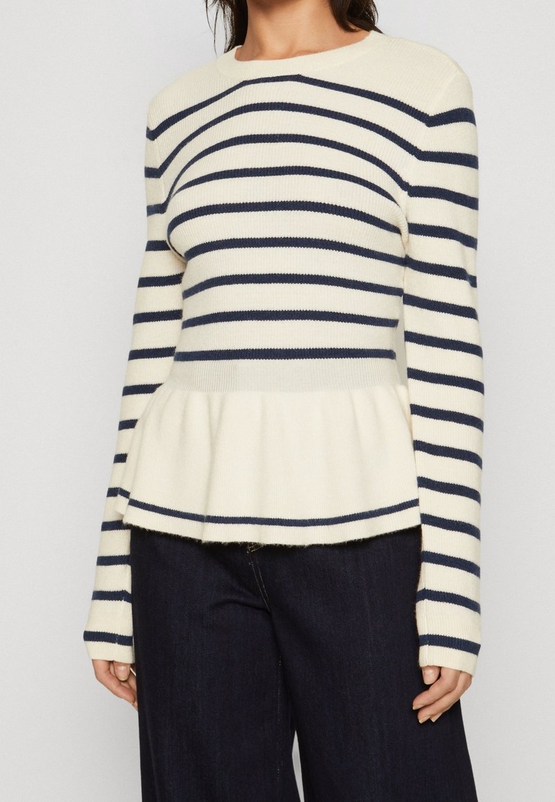 Pull rayé en crème et marine, texture côtelée, col rond, manches longues, ourlet peplum. Associé à un pantalon en denim foncé.