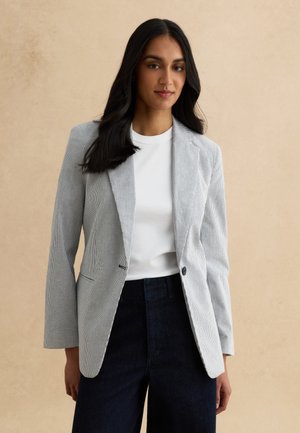 Vrouw met lang donker haar, gekleed in een wit shirt, lichtgrijze blazer met fijne strepen en donkergroene broek met hoge taille, staande tegen een beige achtergrond.