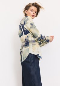 Button-up shirt in soft fabric, featuring a yellow and blue abstract pattern, with a rounded hem, and long sleeves. Paired with dark denim pants.