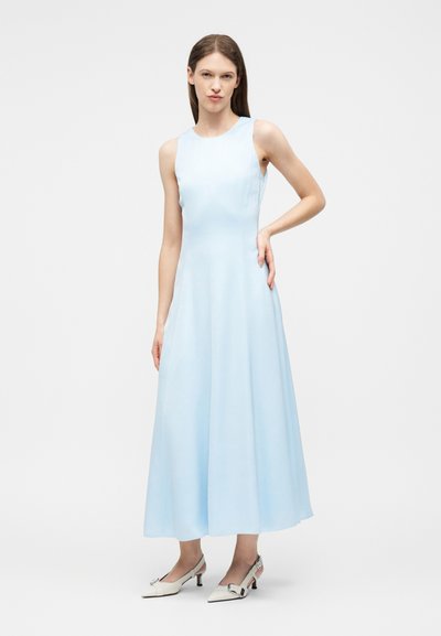 Femme debout portant une robe midi sans manches bleu clair associée à des chaussures blanches à talons bas et bride arrière, sur un fond uni.