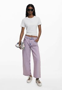 Weißes tailliertes T-Shirt, lavendelfarbene Jeans mit weitem Bein und weiße Turnschuhe. Accessoires sind eine mehrfarbige gemusterte Tasche und dunkle Sonnenbrillen.