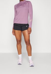 Henkilö, jolla on päällään violetti pitkähihainen urheilu-paita, mustat shortsit, valkoiset sukat ja vaalean violetit juoksukengät, seisoo yksinkertaisen taustan edessä.