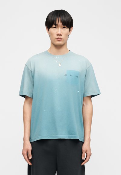 Νέος άνδρας με ίσια μαύρα μαλλιά που φοράει ανοιχτό μπλε φαρδιά μπλούζα με μικρό λογότυπο "mbh" στο στήθος και μαύρο παντελόνι.