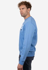 Sudadera azul claro con cuello redondo, puños y dobladillo acanalados. Presenta rayas gráficas en diferentes tonos de azul y blanco a lo largo del pecho.