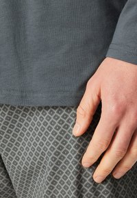 Graues Langarmshirt mit glatter Textur, kombiniert mit gemusterten grauen Hosen, die ein geometrisches Rautenmuster aufweisen. Hand ruht auf der Hose.