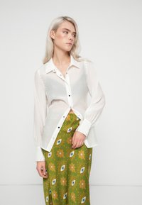Mulher com cabelos loiros longos, vestindo uma blusa branca transparente com botões e uma saia verde com um padrão de olhos e flores por toda parte.