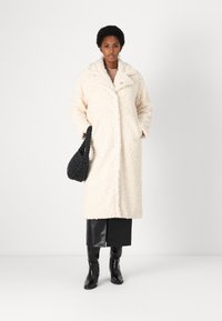 Crème kleurige, textuur coat met een ingeknipt kraag en knoopsluiting. Gepaarde met een zwarte geweven tas en kniehoge laarzen.