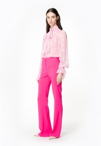 Roze geplooide blouse met een halsband en transparante lange mouwen, gecombineerd met hoge, uitlopende roze broeken en beige puntige schoenen.