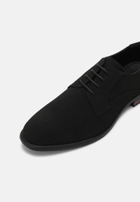 Chaussure noire à lacets en tissu doux, présentant un design épuré, un bout pointu et des détails subtils de couture. Semelle plate avec un accent minimal.