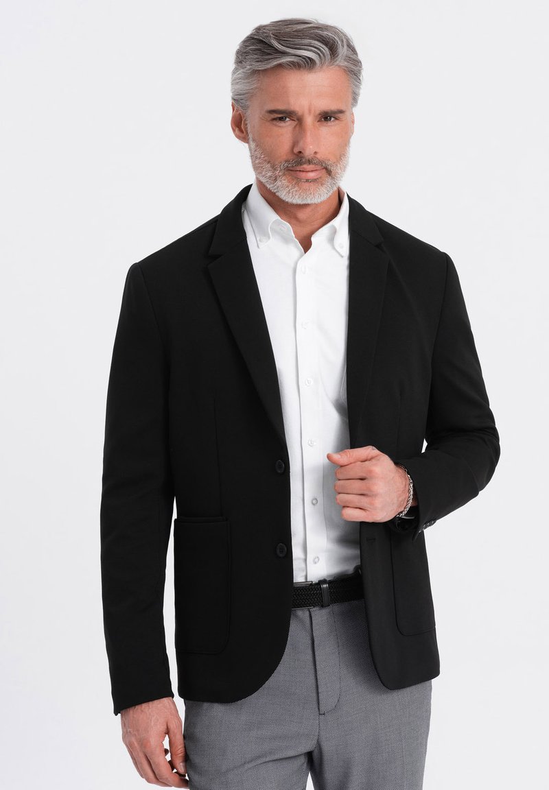 Schwarzes Blazer mit taillierter Passform, einem Knopf, Revers und Vordertaschen, kombiniert mit einem weißen Hemd und hellgrauen gemusterten Hosen.
