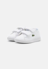 Zapatillas blancas con parte superior sintética lisa, doble correa de Velcro, puntera redondeada y un pequeño logo verde en el costado.