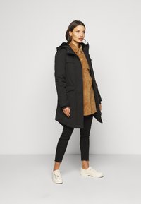 Parka negra con capucha, cierre de botones y dos bolsillos frontales, llevada sobre una camisa de pana marrón y jeans negros; combinada con zapatillas blancas.
