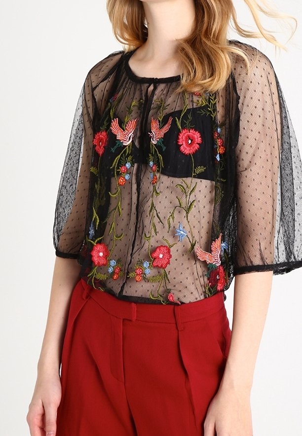 Blouse noire transparente avec fleurs rouges brodées, accents bleus et oiseaux roses, associée à un pantalon rouge taille haute.