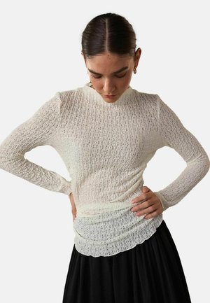 Weißes langärmliges Oberteil mit strukturiertem, gemustertem Stoff und einem gewellten Saum, kombiniert mit einem schwarzen Rock. Verfügt über eine enganliegende Silhouette.