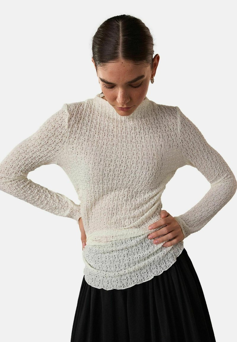 White long-sleeve top with textured, patterned fabric and a scalloped hem, paired with a black skirt. Features a fitted silhouette.