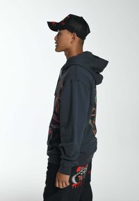 Mørkegrå hættetrøje med livlige multicolore grafikker på siderne. Sammen med sorte denimjeans med markante broderede detaljer. Sort mesh-cap.