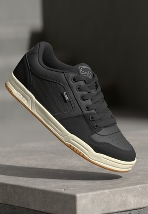 Chaussure de skate noire avec semelle blanche et semelle extérieure en gomme, arborant la marque "Globe" sur l'étiquette latérale, la languette et la semelle intermédiaire, flottant au-dessus d'une surface en béton.