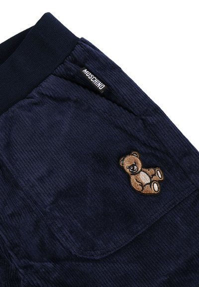 Pantaloni di velluto a coste blu navy con vita elasticizzata, caratterizzati da una toppa di un orsetto marrone e un'etichetta nera del marchio Moschino vicino alla tasca.