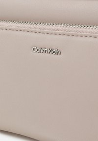 Borsa in ecopelle beige con inserti in metallo argentato, dotata di una zip visibile e dettaglio in rilievo del logo "Calvin Klein" sulla parte anteriore.