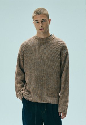 Jeune homme aux cheveux courts portant un pull en maille brun et un pantalon foncé, debout avec une main dans la poche sur un fond uni.