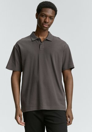 Polo shirt gris foncé avec un design col, manches courtes, patte de boutonnage à deux boutons et un motif texturé subtil. Associé à un pantalon noir.