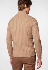 Beżowy sweter w sploty z wysokim kołnierzem, ściągaczami u dołu i przy rękawach, wzorzysty na całym ciele, noszony z brązowymi spodniami na neutralnym tle.