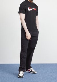 Svart t-shirt med vitt och rött "AIR"-logotyp, kombinerad med svarta byxor och svart-vita sneakers med röda detaljer. Enkelt, avslappnat design.