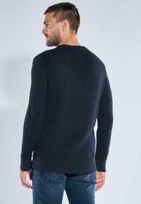 Mann mit kurzen braunen Haaren und Bart, der einen dunkelblauen Strickpullover und blaue Jeans trägt, seitlich gewandt, sodass sein Rücken größtenteils zu sehen ist.
