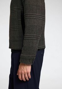 Veste en laine à carreaux gris foncé avec une finition texturée, dotée d'un poignet boutonné, portée sur un pantalon marine, vue de côté.