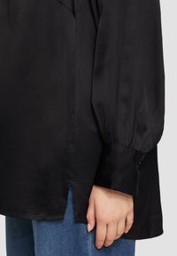 Zwarte satijnen blouse met pofmouwen, zijsplitten aan de onderkant en een zachte textuur. Zichtbare hand die de stof vasthoudt nabij denim jeans.