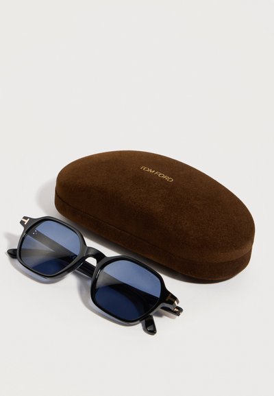 Čierne okuliare so štvorcovými rámami a modrými šošovkami, ktoré ležia vedľa hnedého pevného puzdra s nápisom "Tom Ford" zlatým písmom.