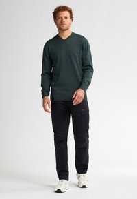 Jersey de punto de color verde oscuro, con cuello en V y mangas largas, combinado con pantalones cargo negros y zapatillas deportivas blancas. Diseño simple con un acabado texturizado.