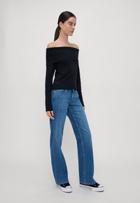 Svart avlang topp med lange ermer, kombinert med lys blå vidde jeans. Modellen har på seg svarte joggesko med hvite såler. Nøytral bakgrunn.