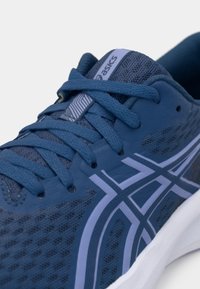 Μπλε αθλητικό παπούτσι με μεσαία πλέξη, αντίθετες ανοιχτό γαλάζιες ρίγες, υφή κορδονιών και έγχυση καουτσούκ στη σόλα. Περιλαμβάνει το λογότυπο ASICS στην γλώσσα.