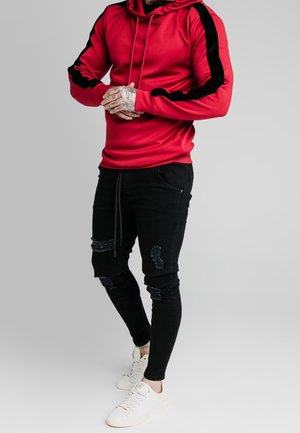 Sweatshirt à capuche rouge avec des rayures noires sur les côtés, porté avec un jean skinny noir déchiré et des baskets blanches. Texture lisse et design ajusté.