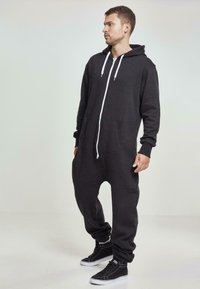 Mono negro de corte holgado con capucha, cremallera blanca y bolsillos frontales. Combinado con zapatillas negras de caña alta. Tejido suave, diseño casual.